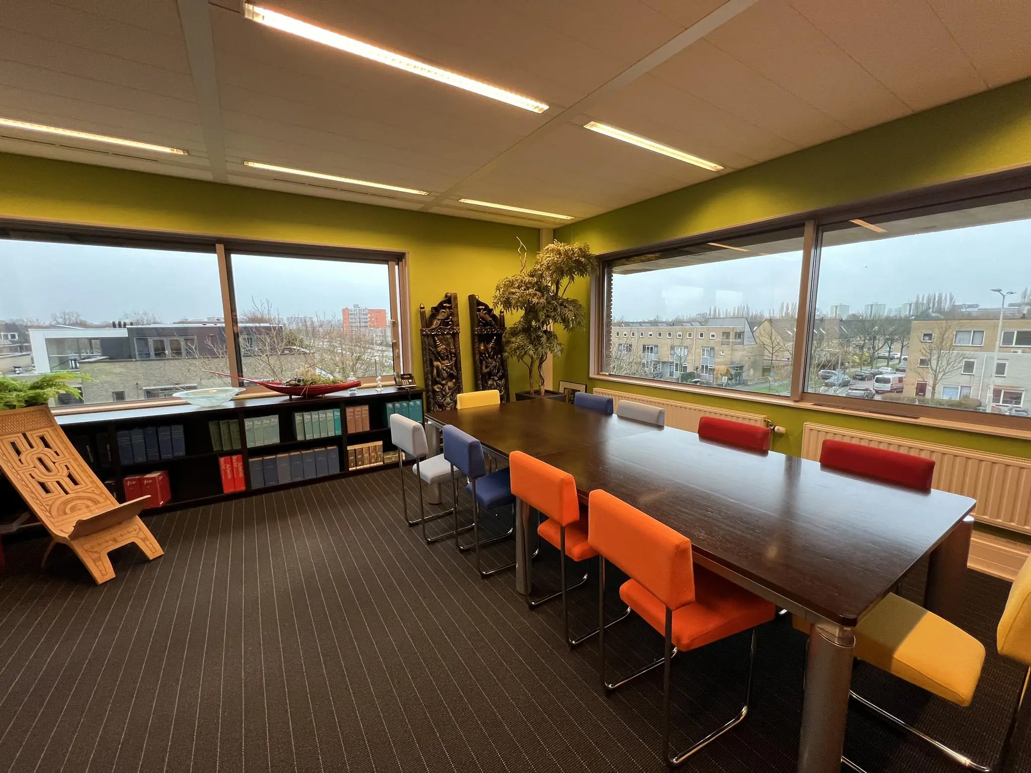 Vergaderruimte aan de Jacques Dutilhweg met kleurrijke stoelen, een lange tafel en uitzicht op een woonwijk.